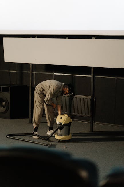 A person in a beige jumpsuit is engaged in surface cleaning using a vacuum cleaner on a carpeted floor within a commercial space. The room features dark wall panels, a large whiteboard or screen mounted on the wall, and a black speaker placed on the floor. The individual is bent over, focusing on cleaning, with natural light illuminating the area, creating a clean and tidy environment. This scene exemplifies professional deep cleaning and sanitisation services by Carpet Cleaners Pimlico, designed for maintaining hygiene and cleanliness in residential or commercial settings.
