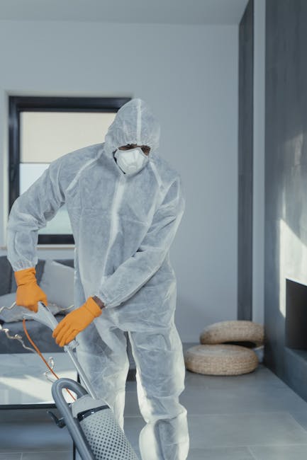 A person dressed in white protective coveralls, a face mask, and orange gloves is operating a vacuum or carpet cleaning machine on a tiled floor within a modern room illuminated by natural light from large windows. The room has a minimalistic design with light-colored walls, and three round cushions are placed against the wall near the window. The scene reflects professional deep cleaning or sanitisation processes aimed at maintaining high hygiene standards, aligning with services offered by Carpet Cleaners Pimlico for surface and carpet cleaning within residential or commercial settings.