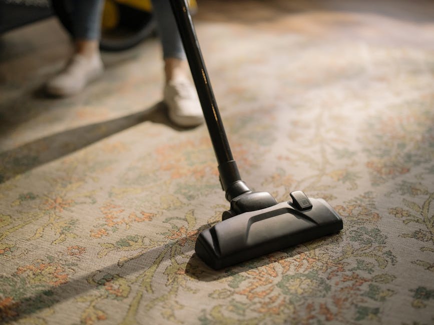 A person in a beige jumpsuit is engaged in surface cleaning using a vacuum cleaner on a carpeted floor within a commercial space. The room features dark wall panels, a large whiteboard or screen mounted on the wall, and a black speaker placed on the floor. The individual is bent over, focusing on cleaning, with natural light illuminating the area, creating a clean and tidy environment. This scene exemplifies professional deep cleaning and sanitisation services by Carpet Cleaners Pimlico, designed for maintaining hygiene and cleanliness in residential or commercial settings.
