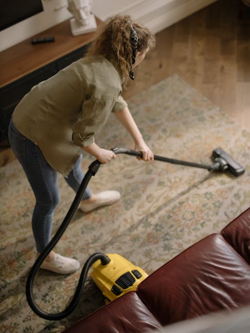 A woman cleaning a patterned area rug with a vacuum cleaner in a living room, which has wooden flooring, a brown leather sofa, and a wooden console table. She is dressed in casual clothing with a beige jacket and jeans, and is focused on surface cleaning. The room is well-lit, with natural light reflecting off the shiny, clean surfaces, demonstrating a deep-cleaning process as part of domestic cleaning services offered by Carpet Cleaners Pimlico. The vacuum's hose is extended towards the rug, ensuring effective sanitisation of the surface. The overall setting emphasizes cleanliness and hygiene within a home environment.