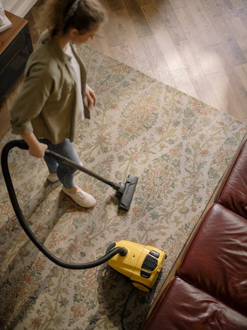 A person kneeling on a patterned area rug in a domestic setting, preparing a yellow and black carpet cleaning device. The individual is wearing a beige jacket and blue jeans, and is opening the device's lid to insert or remove cleaning accessories. The scene shows a close-up of the venue's carpet surface, which appears clean and well-maintained, with soft lighting highlighting the texture of the rug. The background features a wooden floor and part of a wall, indicating a living room or similar indoor space. This image exemplifies surface cleaning and deep cleaning techniques used by Carpet Cleaners Pimlico to maintain hygiene and cleanliness in residential spaces.