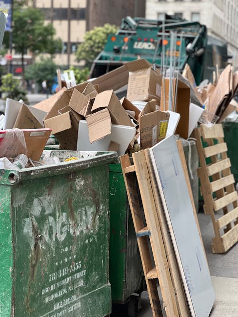 A large green waste bin filled with assorted bulky wet waste, including cardboard boxes, packaging materials, and wooden pallets, situated outdoors on a city street in Pimlico. The background features a street scene with trees, a green waste collection vehicle, and surrounding buildings. The waste appears to be part of a cleanup or renovation process, with some items stacked beside the bin. Natural daylight illuminates the scene, highlighting the various textures and materials of the waste. This image reflects waste management and disposal services offered by Carpet Cleaners Pimlico, especially related to handling bulky wet waste following cleaning or renovation projects. The scene emphasizes proper disposal methods for different types of waste in an urban setting.