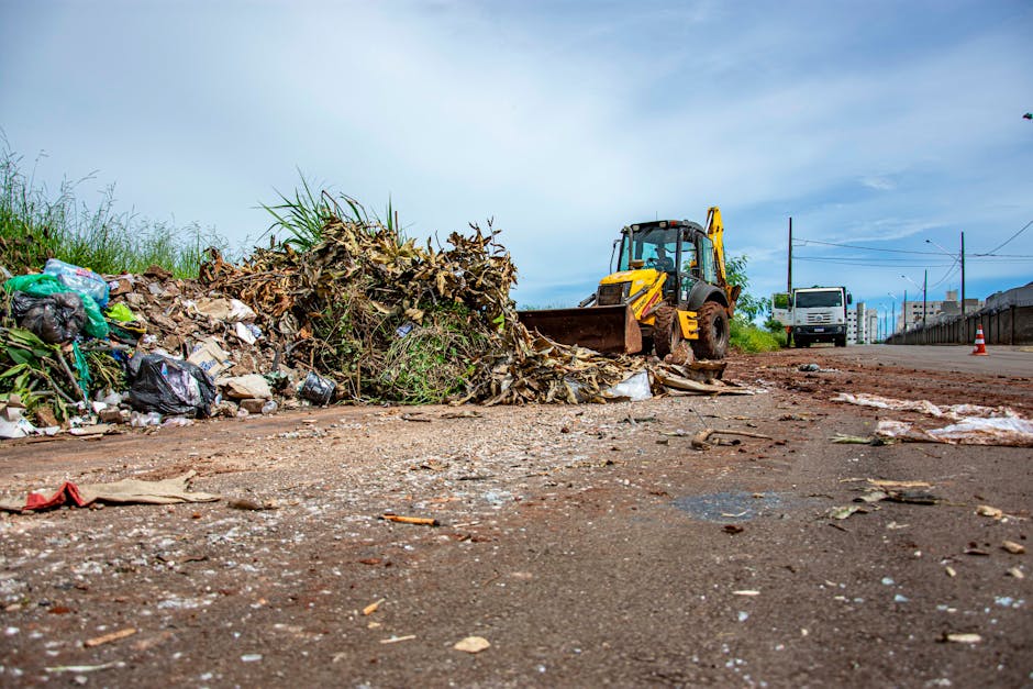 The image depicts a street scene during daytime with a large pile of bulky wet waste and debris including leaves, plastic bags, and miscellaneous refuse accumulated along the edge of the road. A yellow backhoe loader is actively engaged in clearing or moving the waste, positioned centrally in the frame with its front bucket lowered near the ground. The surface of the road appears muddy and scattered with litter, indicating recent waste removal activity. The surroundings feature grassy areas with leafy plants and an overcast sky overhead, with a few vehicles and power lines visible in the background. The scene highlights the importance of effective waste management and sanitation efforts, as associated with professional cleaning and waste removal services offered by Carpet Cleaners Pimlico, especially following cleanup operations involving bulky wet waste removal after incidents or site clearances in Pimlico.