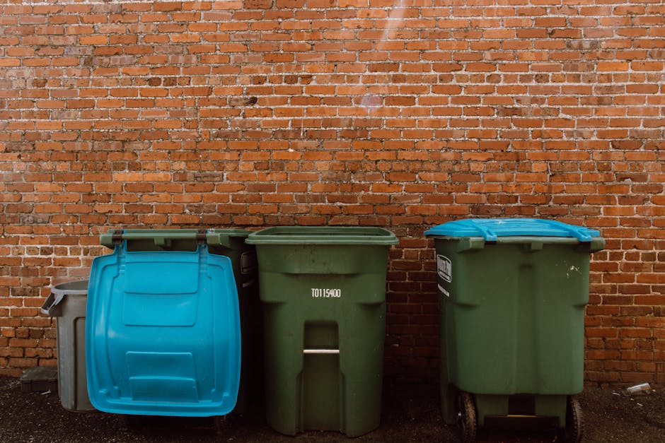 A large green waste bin filled with assorted bulky wet waste, including cardboard boxes, packaging materials, and wooden pallets, situated outdoors on a city street in Pimlico. The background features a street scene with trees, a green waste collection vehicle, and surrounding buildings. The waste appears to be part of a cleanup or renovation process, with some items stacked beside the bin. Natural daylight illuminates the scene, highlighting the various textures and materials of the waste. This image reflects waste management and disposal services offered by Carpet Cleaners Pimlico, especially related to handling bulky wet waste following cleaning or renovation projects. The scene emphasizes proper disposal methods for different types of waste in an urban setting.
