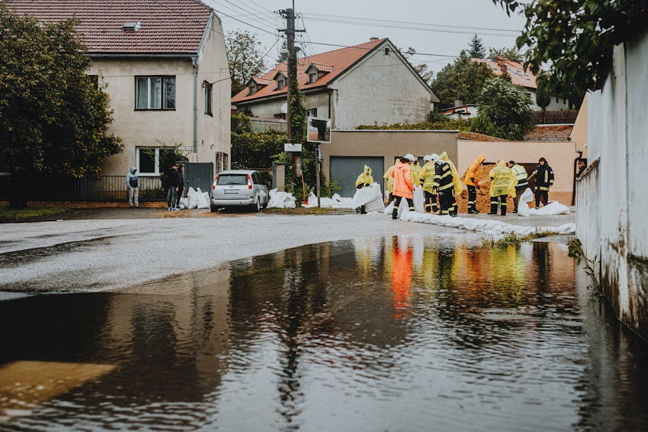 A residential street scene with several workers in high-visibility jackets and protective clothing managing flood response activities. The pavement is wet, and there are large white sandbags stacked along the edge of the road, indicating flood mitigation efforts. The background features suburban houses with tiled roofs and trees, under an overcast sky. The presence of water partially covering the road suggests recent flooding, and the workers are likely involved in cleaning or decontaminating affected surfaces. The area is organized, with equipment and personnel engaged in the recovery process. This image relates to emergency flood response and surface cleaning services provided by Carpet Cleaners Pimlico, emphasizing swift action and thorough sanitisation of residential environments after flooding.