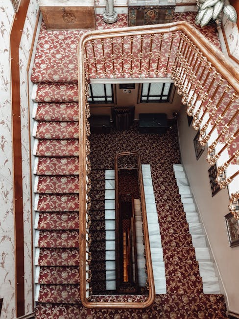 View of a multi-story staircase taken from the upper floor, featuring ornate wooden handrails and balustrades with gold accents. The staircase is covered with a patterned red and beige carpet, which appears clean and well-maintained. The walls adjacent to the staircase display framed artwork, and the lighting is soft, highlighting the detailed design of the interior. The image demonstrates the importance of surface cleaning and maintenance for stairs and handrails in residential or commercial properties, aligning with the services offered by Carpet Cleaners Pimlico, as referenced on their webpage about legal obligations and standards for landlord property care.