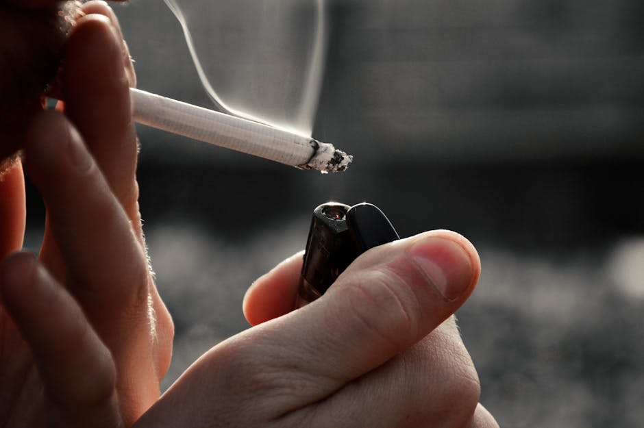 Close-up image showing a person holding a lit cigarette with ash at the tip, positioned near a black lighter held in their other hand. The background appears to be an indoor setting with neutral tones, possibly a living area or kitchen, with soft lighting. The person's fingers are visible, with the cigarette resting between their index and middle fingers, and the lighter being gripped securely. The scene emphasizes the act of smoking, which Carpet Cleaners Pimlico can address through professional cleaning and odour removal, especially in residential spaces affected by cigarette smoke and odours.
