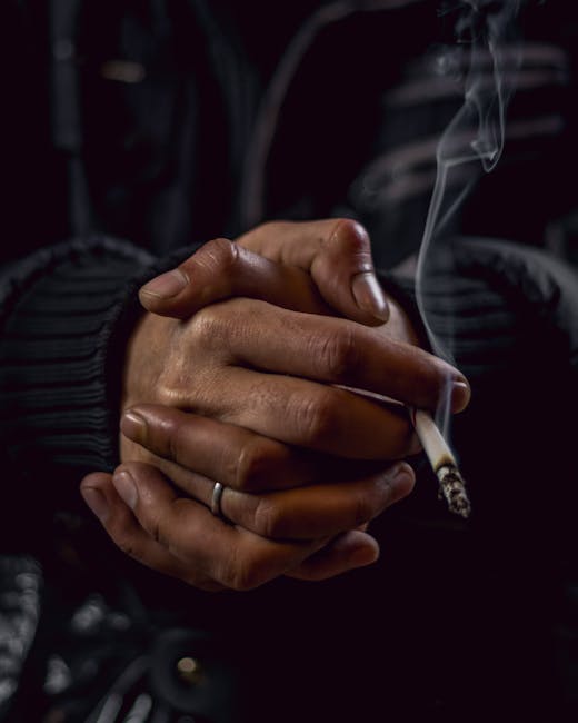 Close-up image of a person's hands clasped together, holding a lit cigarette with visible smoke curling upward. The individual is wearing a dark, textured jacket and a ring on one finger. The background is dark and blurred, highlighting the hands and cigarette which appear slightly oily and stained from cigarette residue. The environment suggests a dimly lit space, possibly indoors, emphasizing the focus on the act of smoking. This visual relates to the topic of cigarette smoke and odour issues, relevant to cleaning and sanitisation services offered by Carpet Cleaners Pimlico for Pimlico flats, as featured on the webpage titled 'Removing cigarette smoke and odours from Pimlico flats, PIMLICO'.