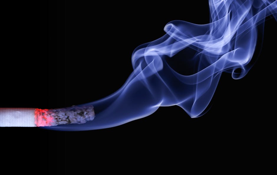 Close-up image showing a person holding a lit cigarette with ash at the tip, positioned near a black lighter held in their other hand. The background appears to be an indoor setting with neutral tones, possibly a living area or kitchen, with soft lighting. The person's fingers are visible, with the cigarette resting between their index and middle fingers, and the lighter being gripped securely. The scene emphasizes the act of smoking, which Carpet Cleaners Pimlico can address through professional cleaning and odour removal, especially in residential spaces affected by cigarette smoke and odours.