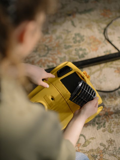 A woman vacuuming a patterned area rug in a living room with wooden flooring. She is holding a yellow vacuum cleaner with a black hose and nozzle, wearing a beige jacket, blue jeans, and white sneaker