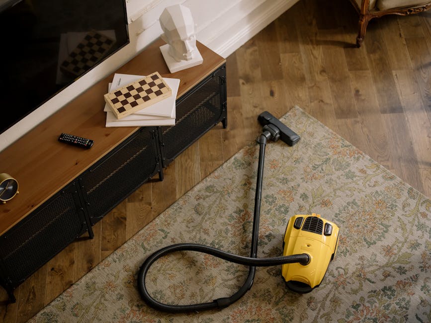 A yellow vacuum cleaner with a long hose attachment is positioned on a patterned area rug in a residential living room. The room features a wooden floor and a black metal TV stand against the wall, which holds objects including a remote control, a white bust sculpture, and some books. The lighting is natural, highlighting the clean and tidy state of the space, indicative of professional surface cleaning and maintenance by Carpet Cleaners Pimlico, a provider specializing in domestic cleaning services in the Pimlico area.