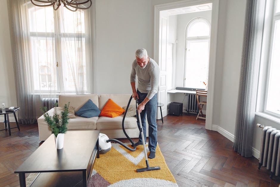 A yellow vacuum cleaner with a long hose attachment is positioned on a patterned area rug in a residential living room. The room features a wooden floor and a black metal TV stand against the wall, which holds objects including a remote control, a white bust sculpture, and some books. The lighting is natural, highlighting the clean and tidy state of the space, indicative of professional surface cleaning and maintenance by Carpet Cleaners Pimlico, a provider specializing in domestic cleaning services in the Pimlico area.